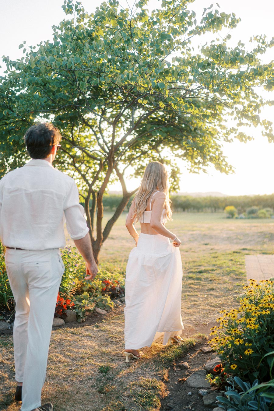 A couple in white attire enjoying a sunny, serene garden setting, with colorful flowers and lush greenery under a bright sunlit sky.