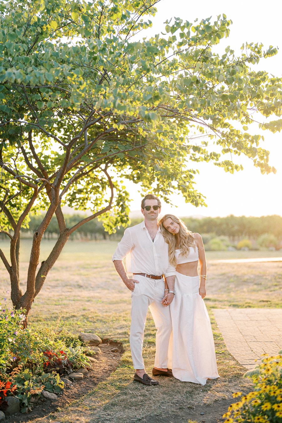 Couple in stylish white attire stands under a tree with lush green leaves at sunset, surrounded by a picturesque garden setting.