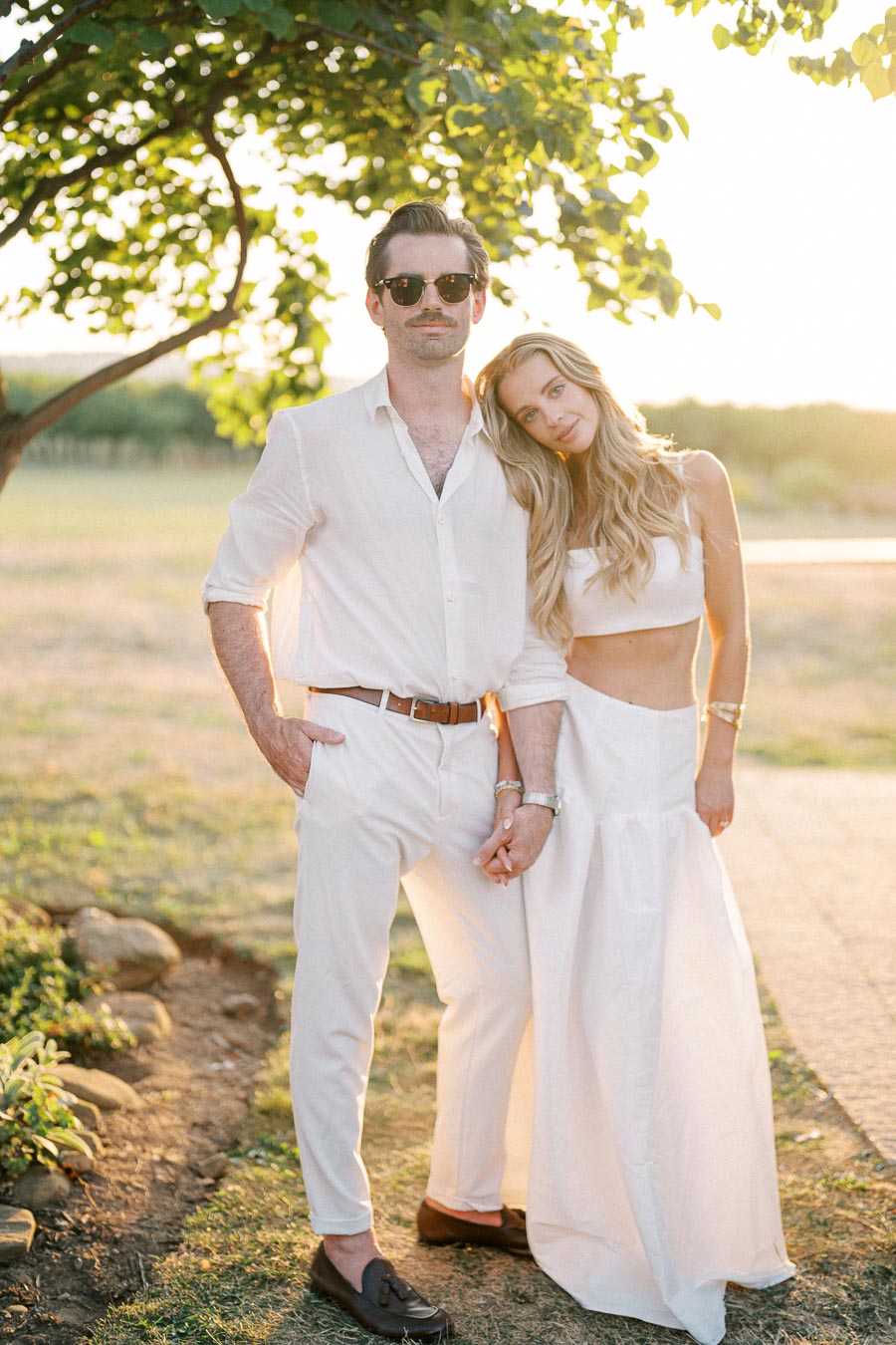 Stylish couple in white outfits posing outdoors at sunset, under a tree in a serene landscape setting