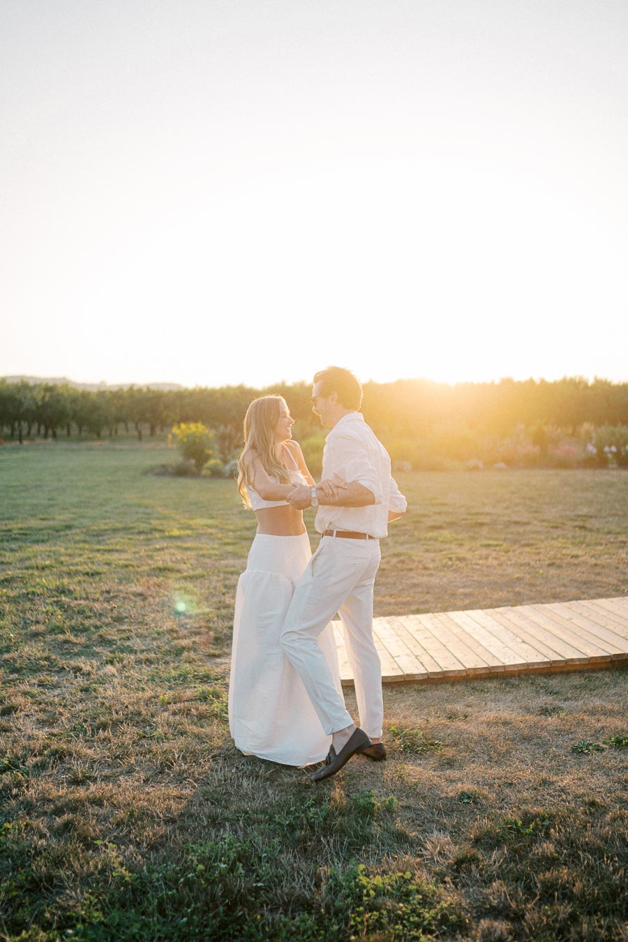 A couple dancing in a scenic outdoor setting during sunset, wearing elegant white clothing, with a lush green landscape in the background.