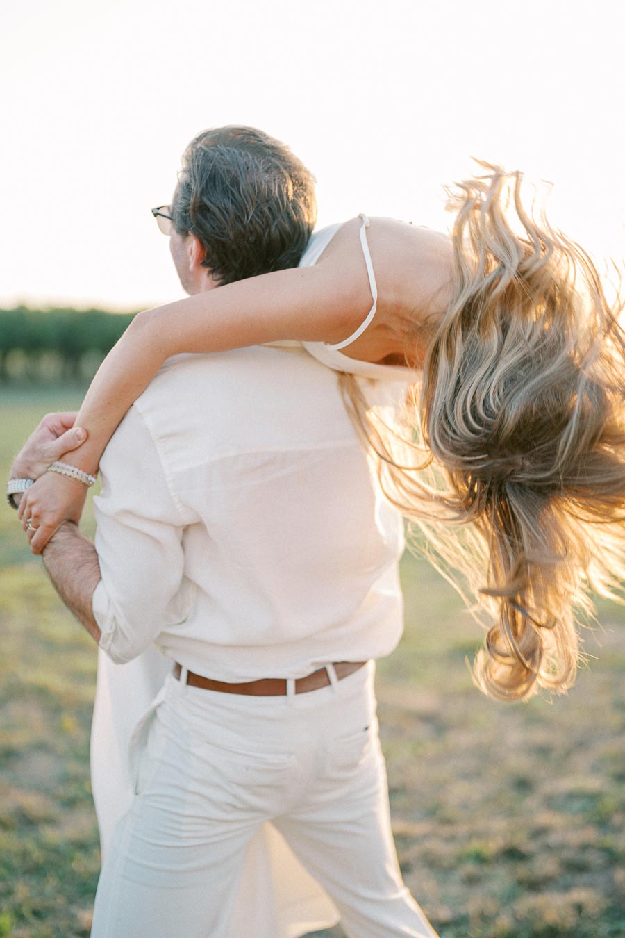 A playful outdoor moment showing a couple dressed in white, with a man carrying a woman over his shoulder in a sunlit field setting.