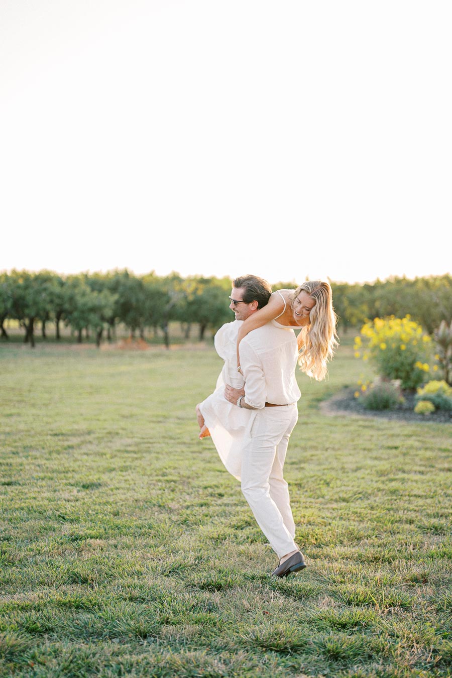 A couple enjoying a playful moment in a sunny, grassy field, with trees and flowers in the background. The man is carrying the woman on his back as they both smile, dressed in light, summery clothes.