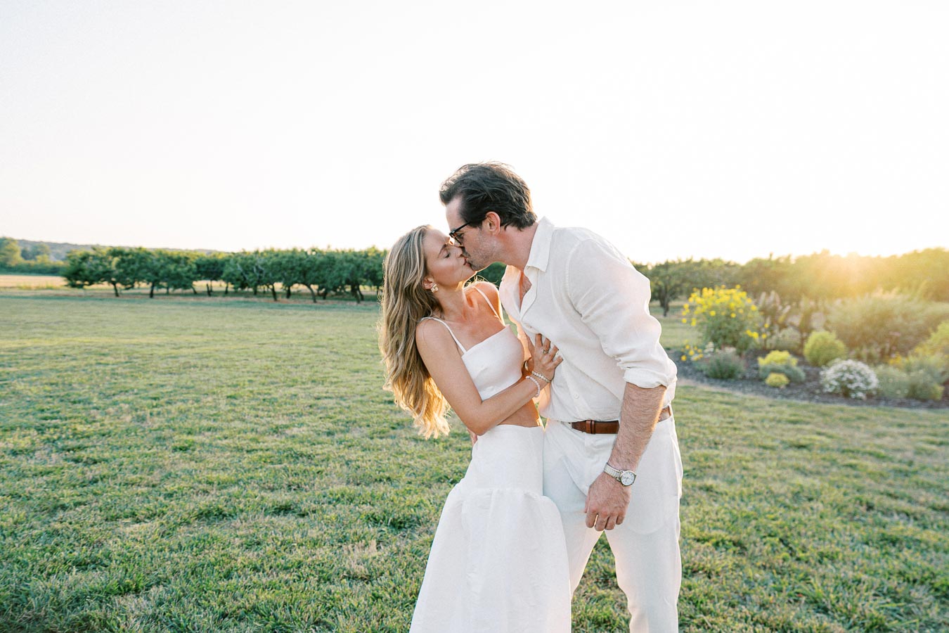 A couple kissing in a sunny outdoor field with lush greenery and flowers, both wearing white outfits, capturing a romantic and serene moment in nature.