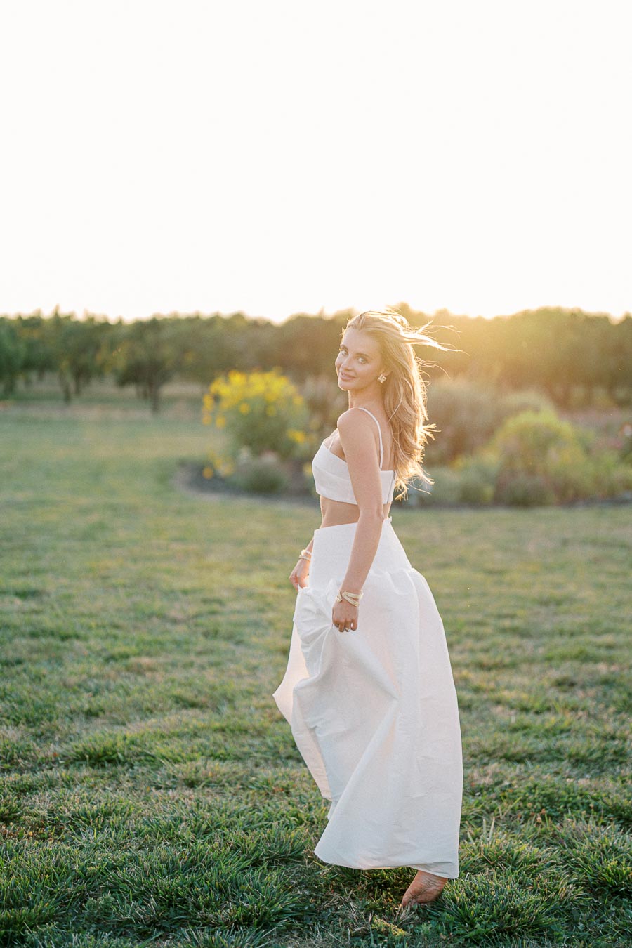 Young woman in a white dress walking through a sunny field with a joyful expression.