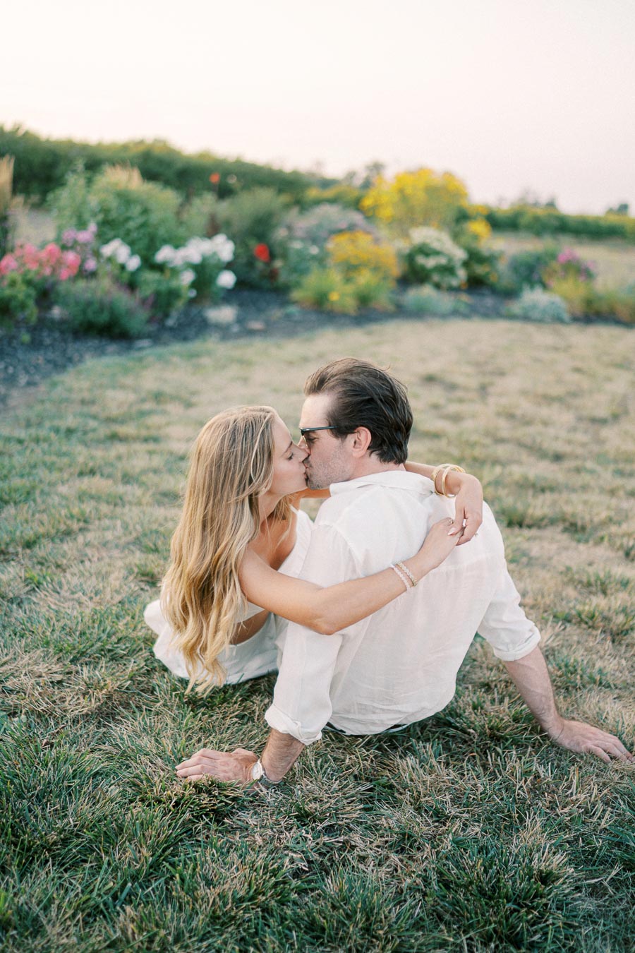 A couple embracing and kissing on a grassy field surrounded by colorful flowers, under a clear sky, conveying romance and tranquility.