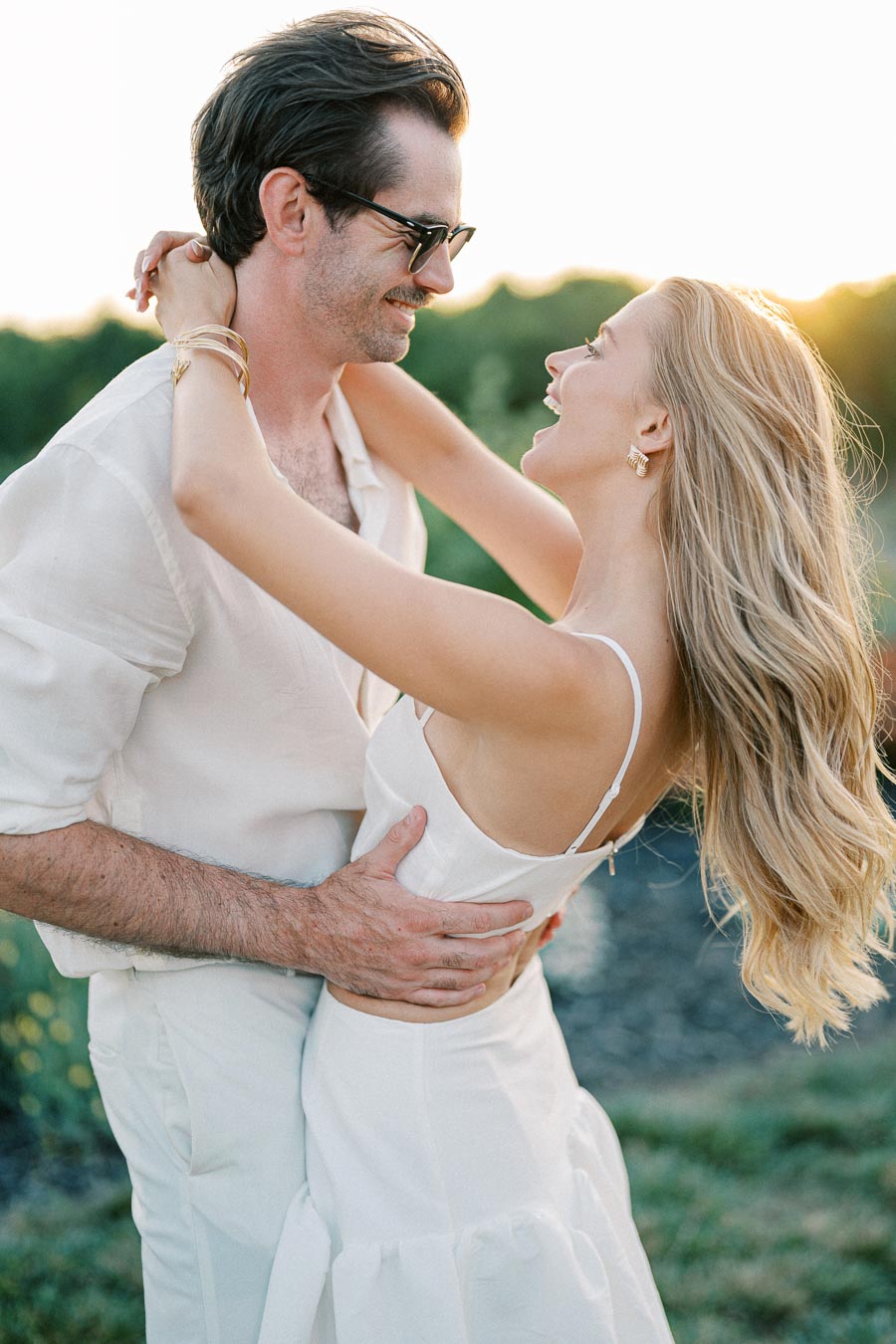A couple embracing and smiling at each other in an outdoor setting during sunset, dressed in white attire, conveying a romantic and joyful moment.