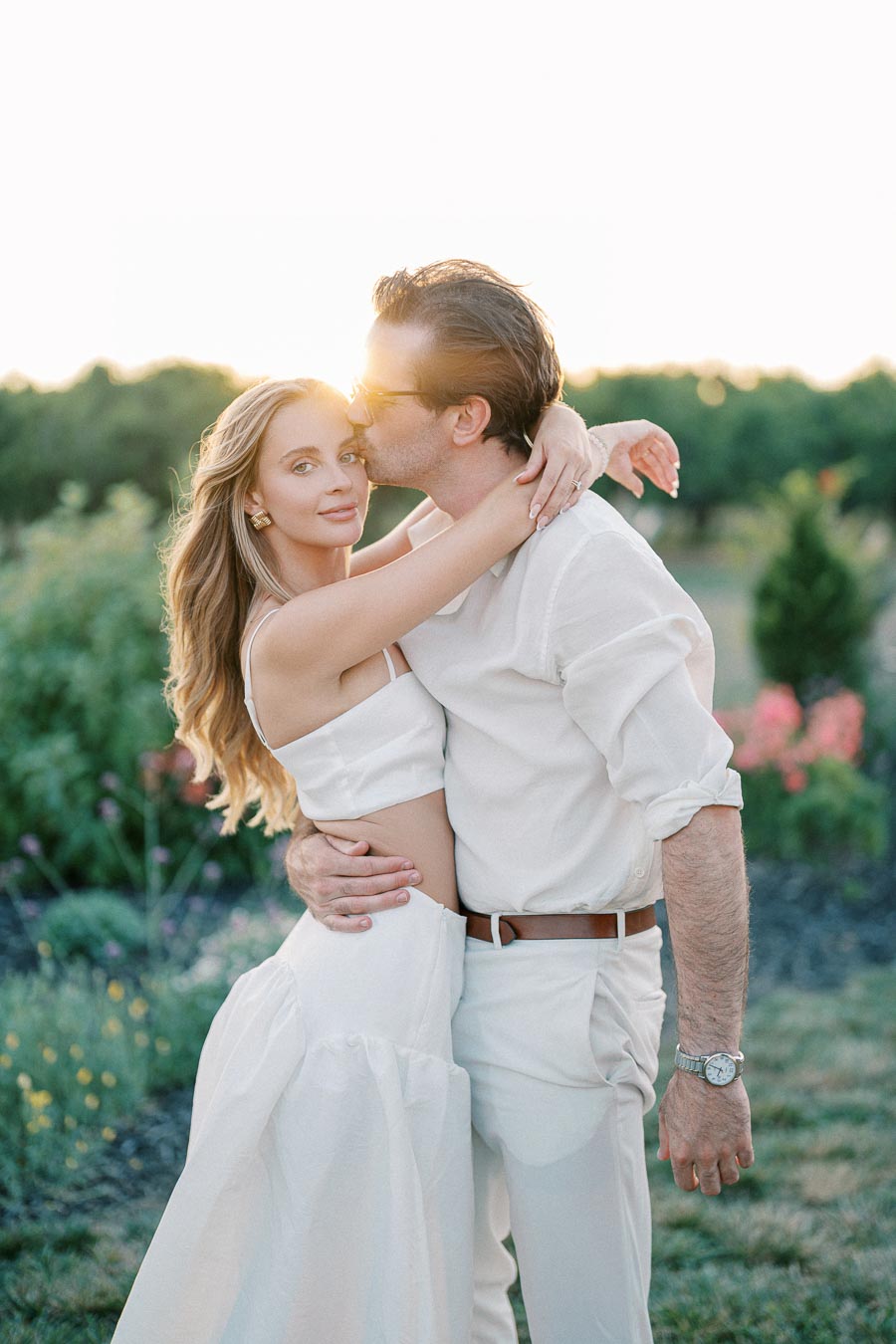 Romantic couple embracing outdoors at sunset, dressed in white, with lush greenery and flowers in the background.