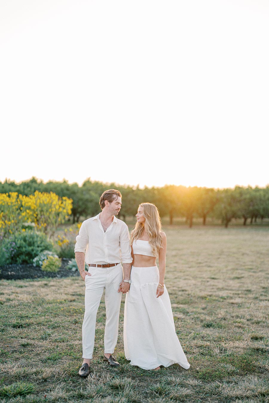 A couple dressed in white holds hands in a sunlit field, with lush greenery and yellow flowers in the background, capturing a serene and romantic moment.