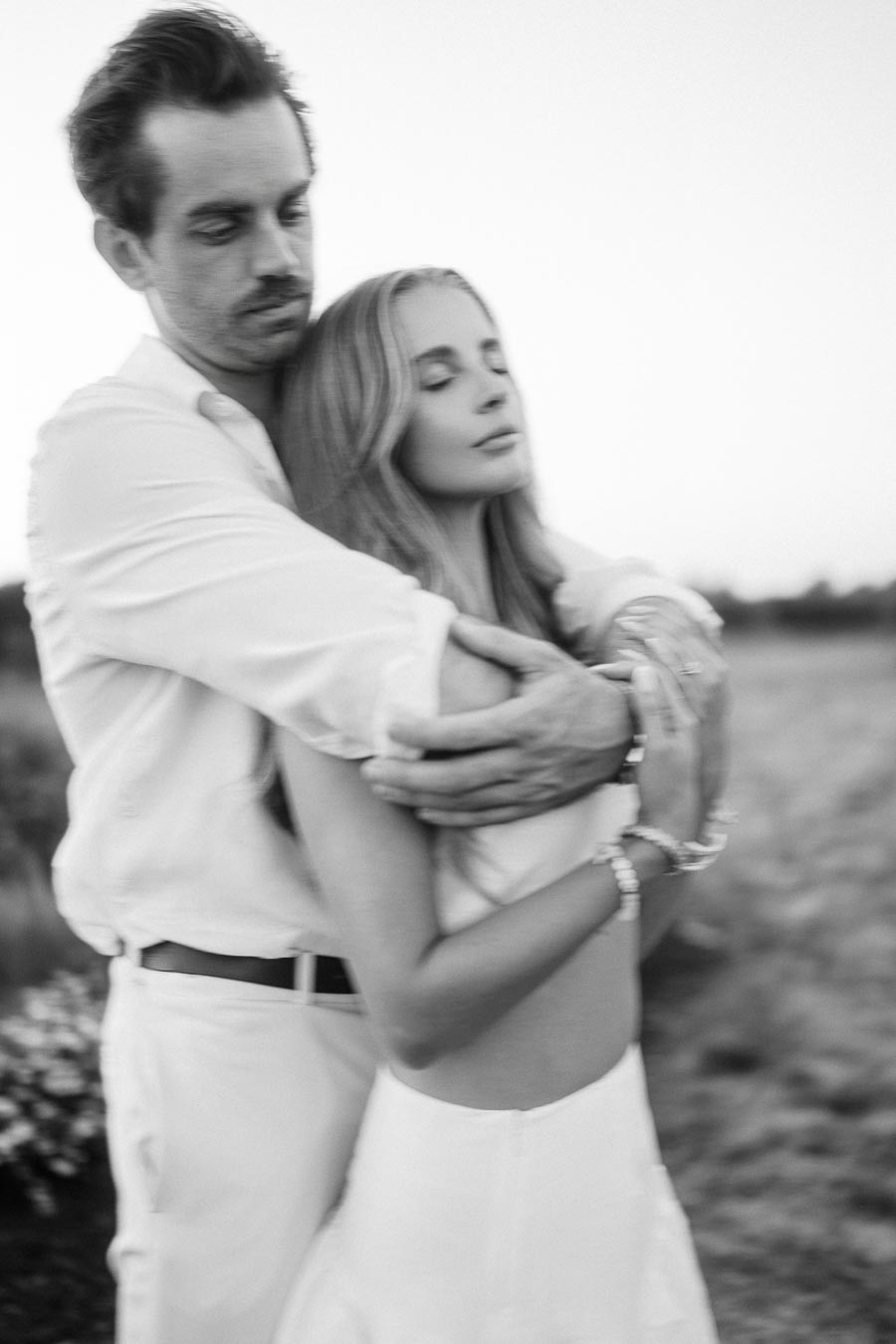 A black and white image of a couple embracing in an outdoor setting, with both wearing light-colored clothing. The woman has long hair and appears to be content with her eyes closed, while the man looks towards the camera. The background is softly blurred with natural scenery.