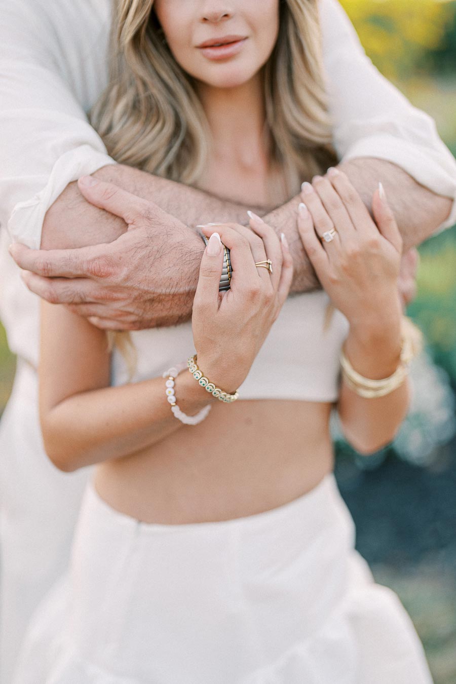 A couple embracing, with the woman's hands adorned with rings and bracelets gently resting on the man's crossed arms, set against a blurred outdoor background.