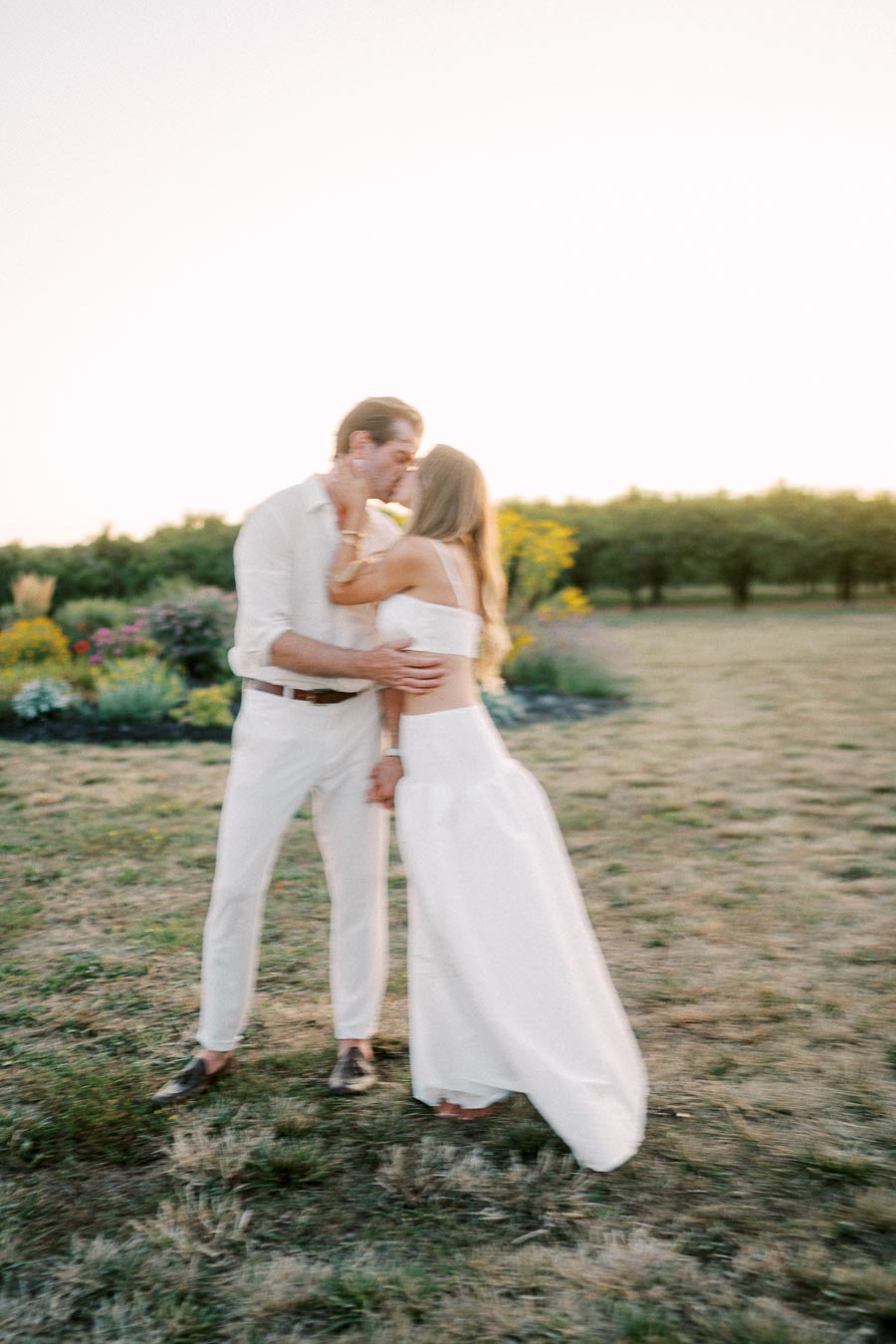 A couple in white attire sharing a romantic kiss in a sunlit field surrounded by flowers and greenery.