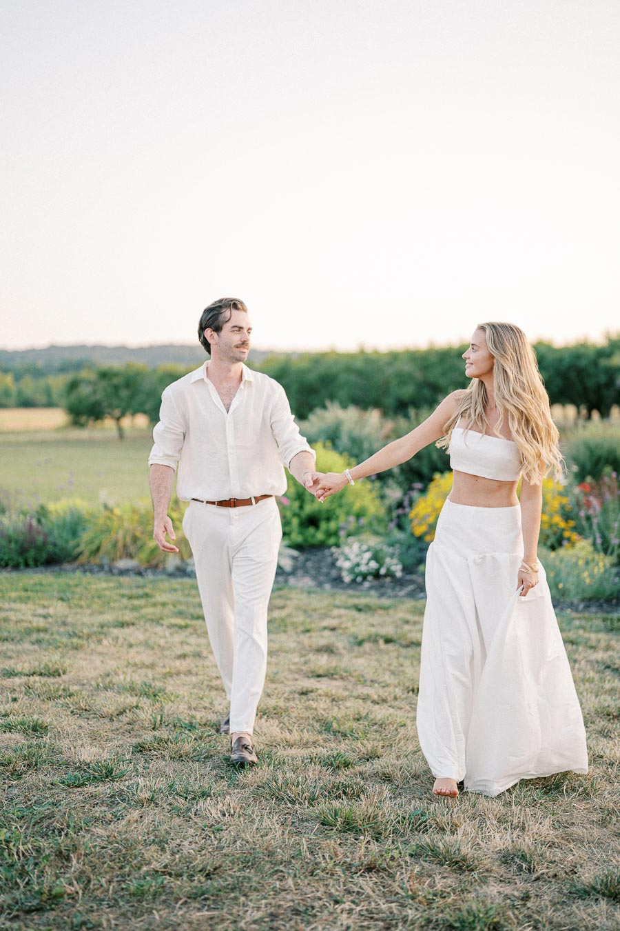 A couple in white attire holding hands and walking through a scenic garden landscape, showcasing a romantic summer setting.
