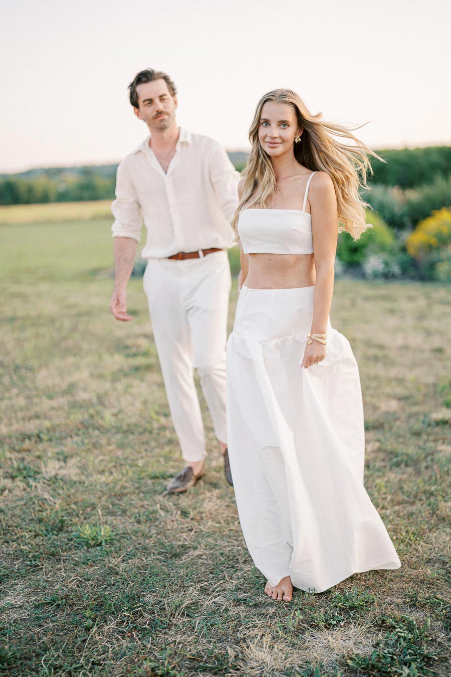 A couple dressed in white outfits walking through a field at sunset, with a serene landscape in the background.