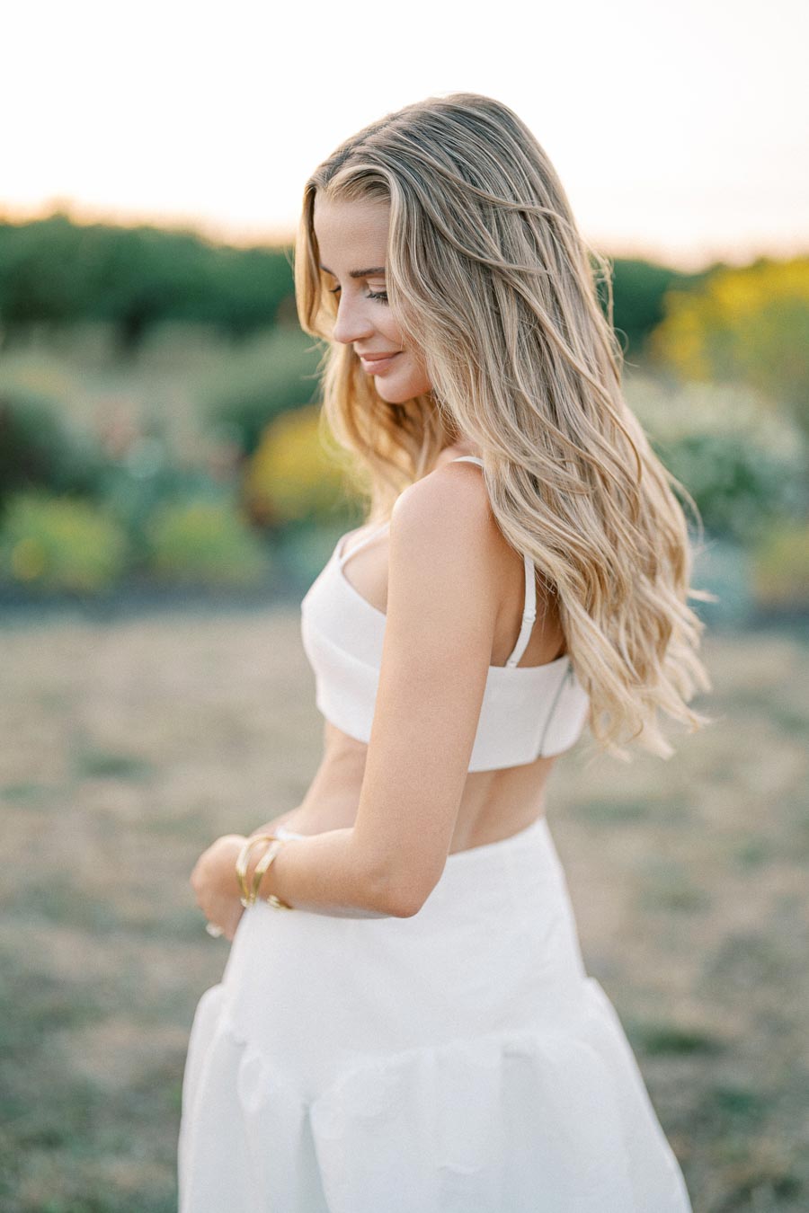 Young woman with long blonde hair wearing a white crop top and skirt, smiling softly and standing in a serene outdoor setting at sunset.
