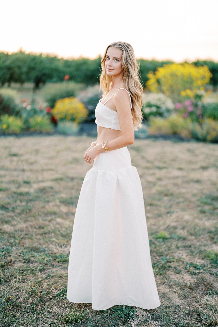 A woman in a white two-piece outfit standing in a grassy garden with colorful flowers and green foliage in the background.
