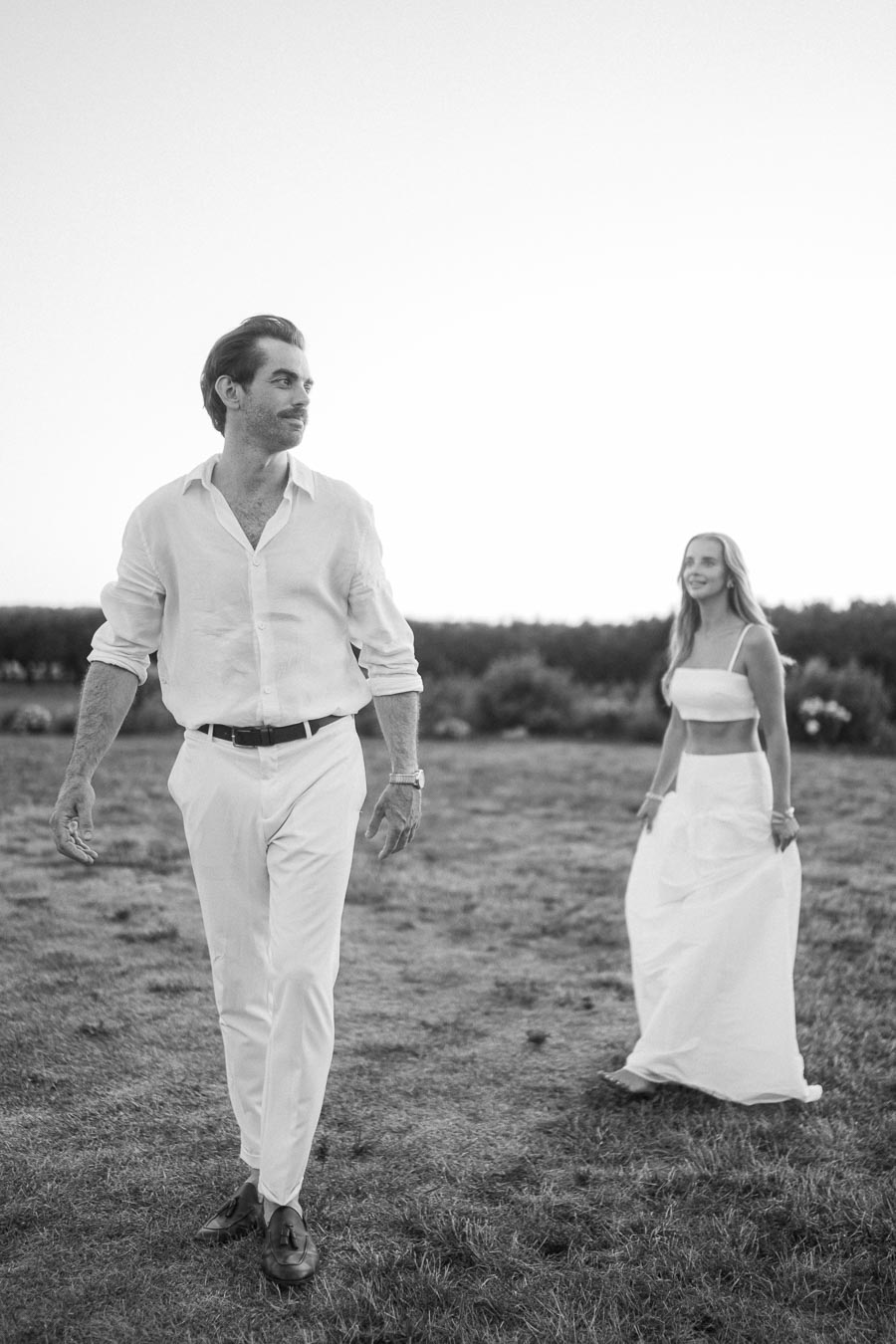 Monochrome image of a man and woman in elegant white attire walking outdoors on grassy terrain, capturing a serene and stylish moment against a clear sky.