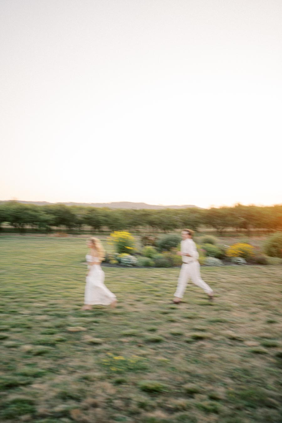 Blurry image of a couple running across a grassy field during sunset, with vibrant flowers in the background.