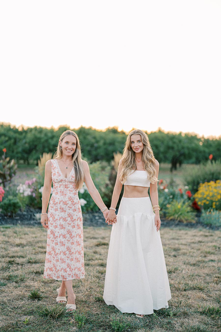 Two women holding hands in a garden setting, one wearing a floral dress and the other in a white skirt and crop top, both smiling at sunset.