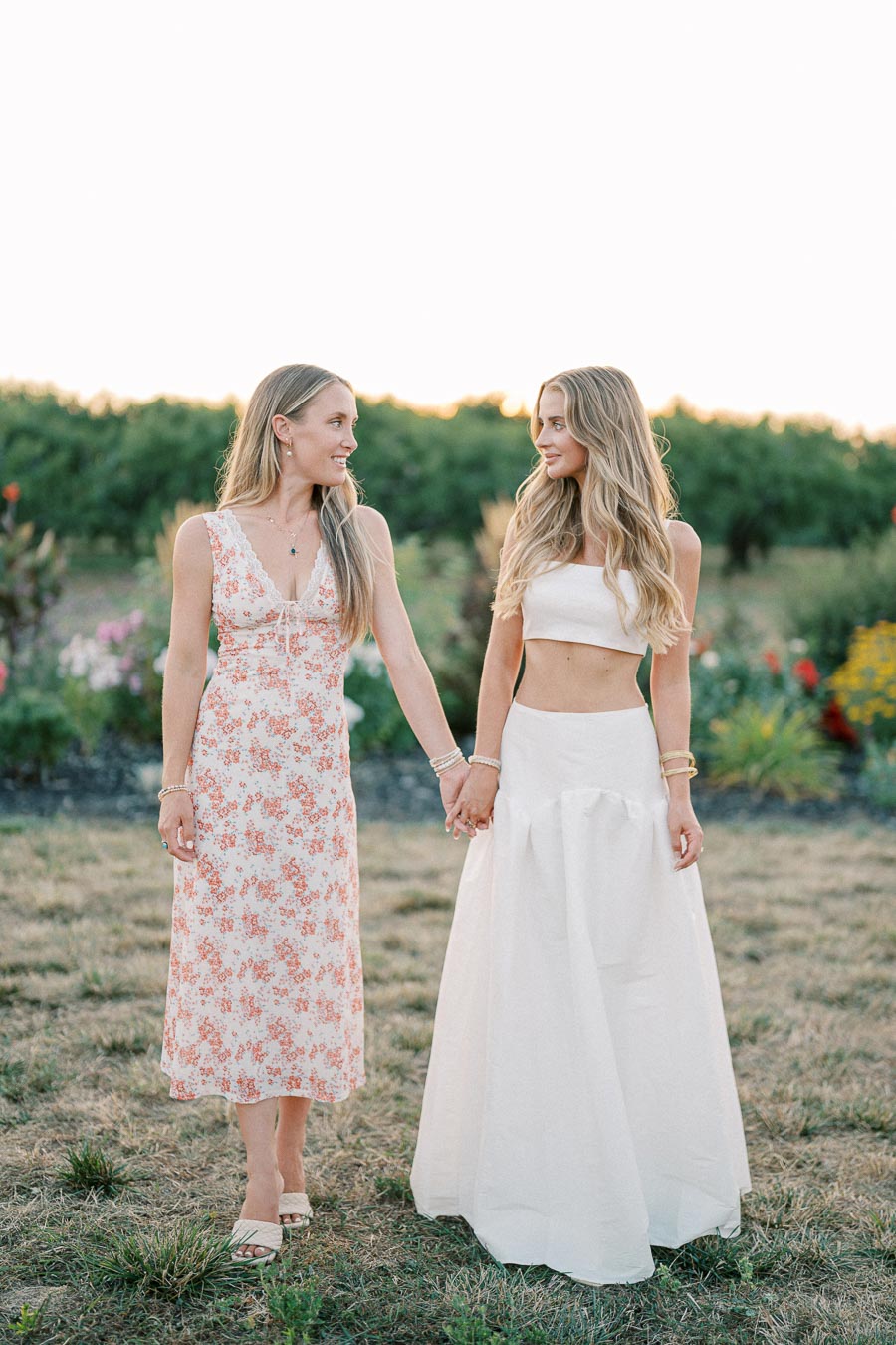 Two women standing hand in hand in a grassy field, one wearing a floral dress and the other in a white two-piece gown, with a blurred natural background of greenery and flowers.