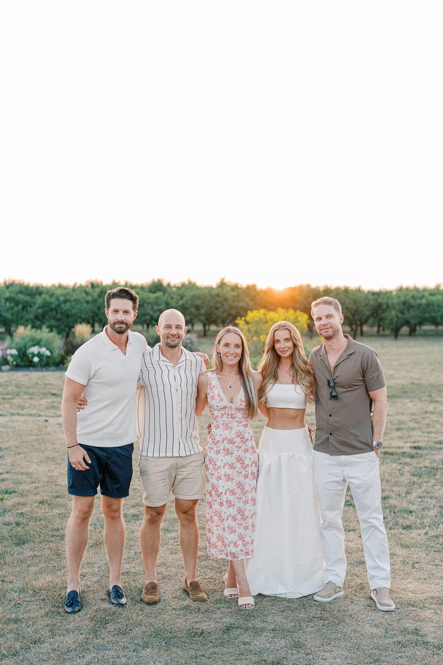 A group of five people posing outdoors at sunset, dressed in summer attire with lush greenery in the background.