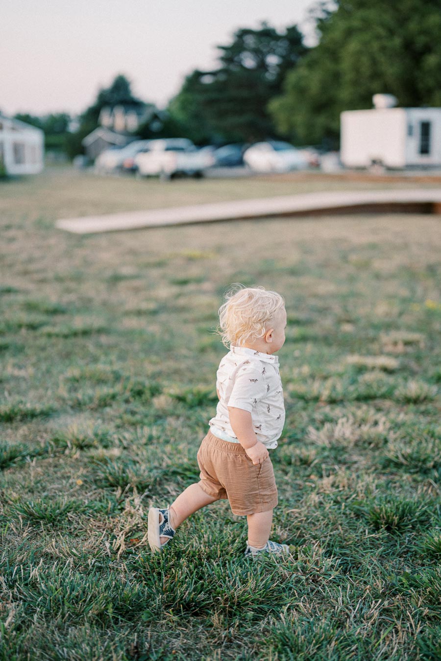 A toddler with blond hair wearing a patterned shirt and brown shorts walking on grass in an outdoor setting with buildings and trees in the background.