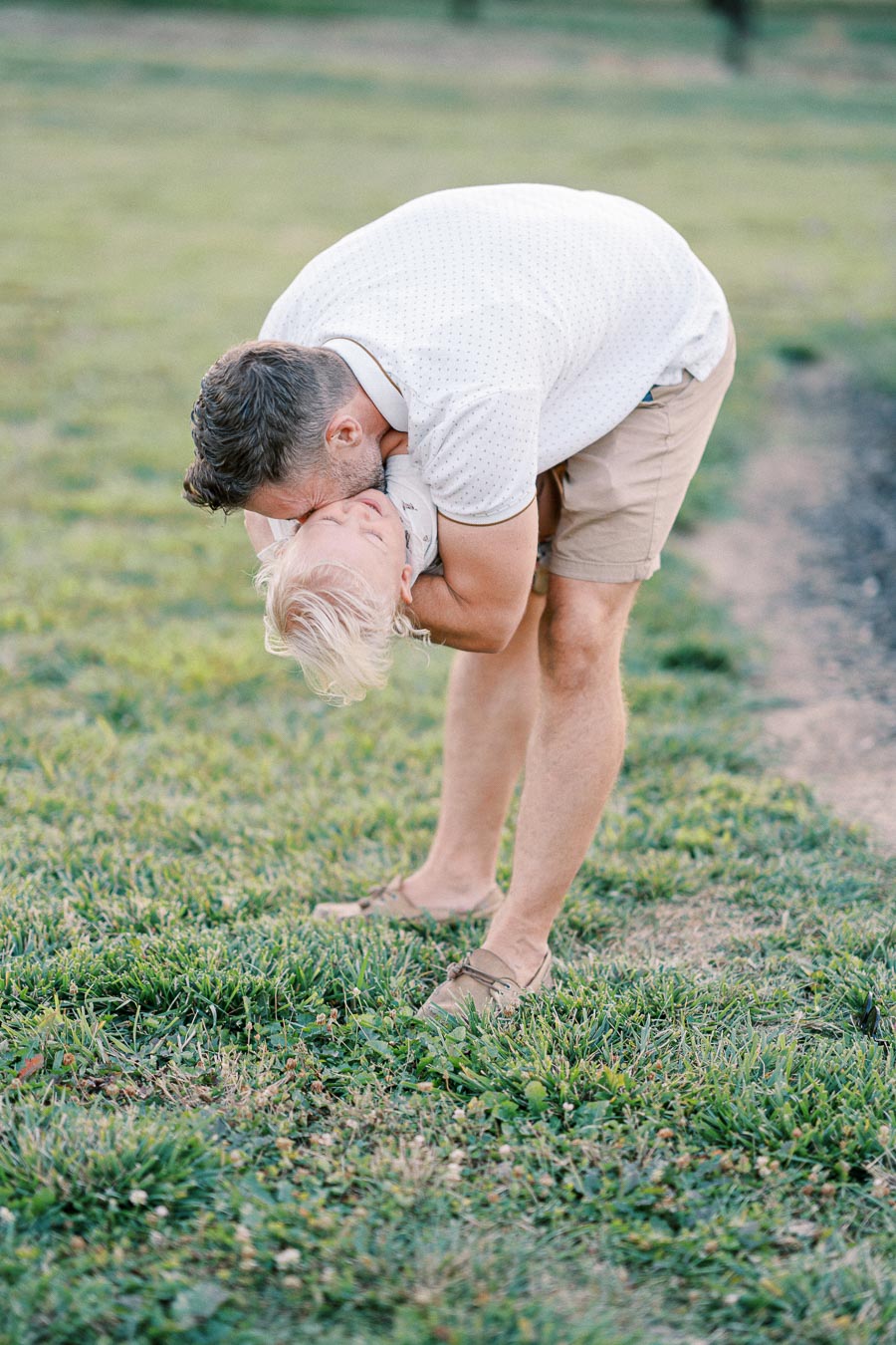 Father playfully holding and kissing his young son upside down on a grassy field, both smiling and enjoying the sunny day.