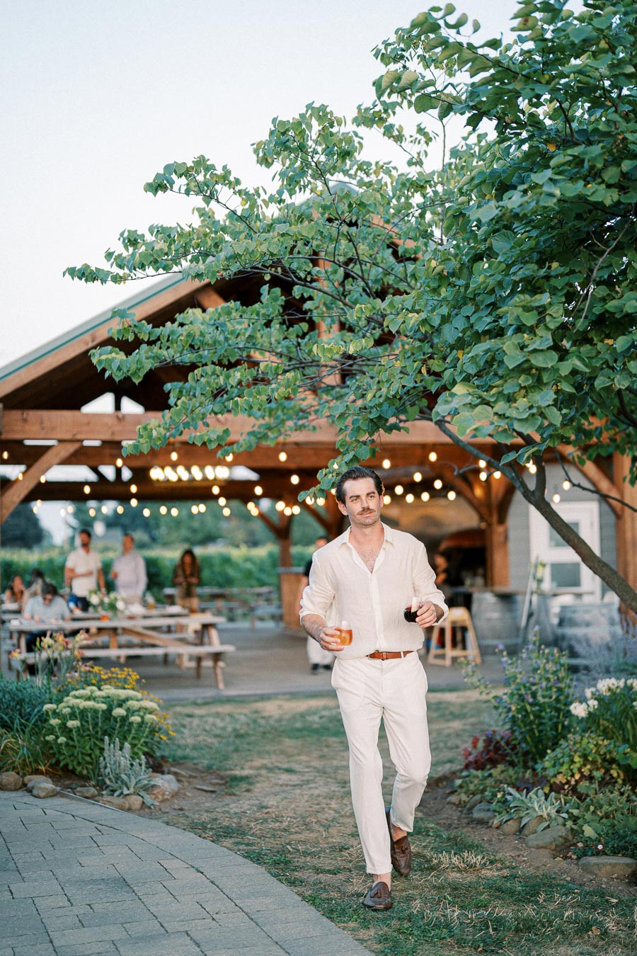 Man in white outfit holding drinks at an outdoor garden party with people and lights in the background.
