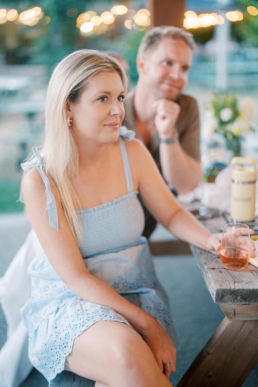 Woman in a light blue sundress holding a glass of rosé wine, sitting at an outdoor wooden table with blurred lights in the background, accompanied by a man leaning on the table.