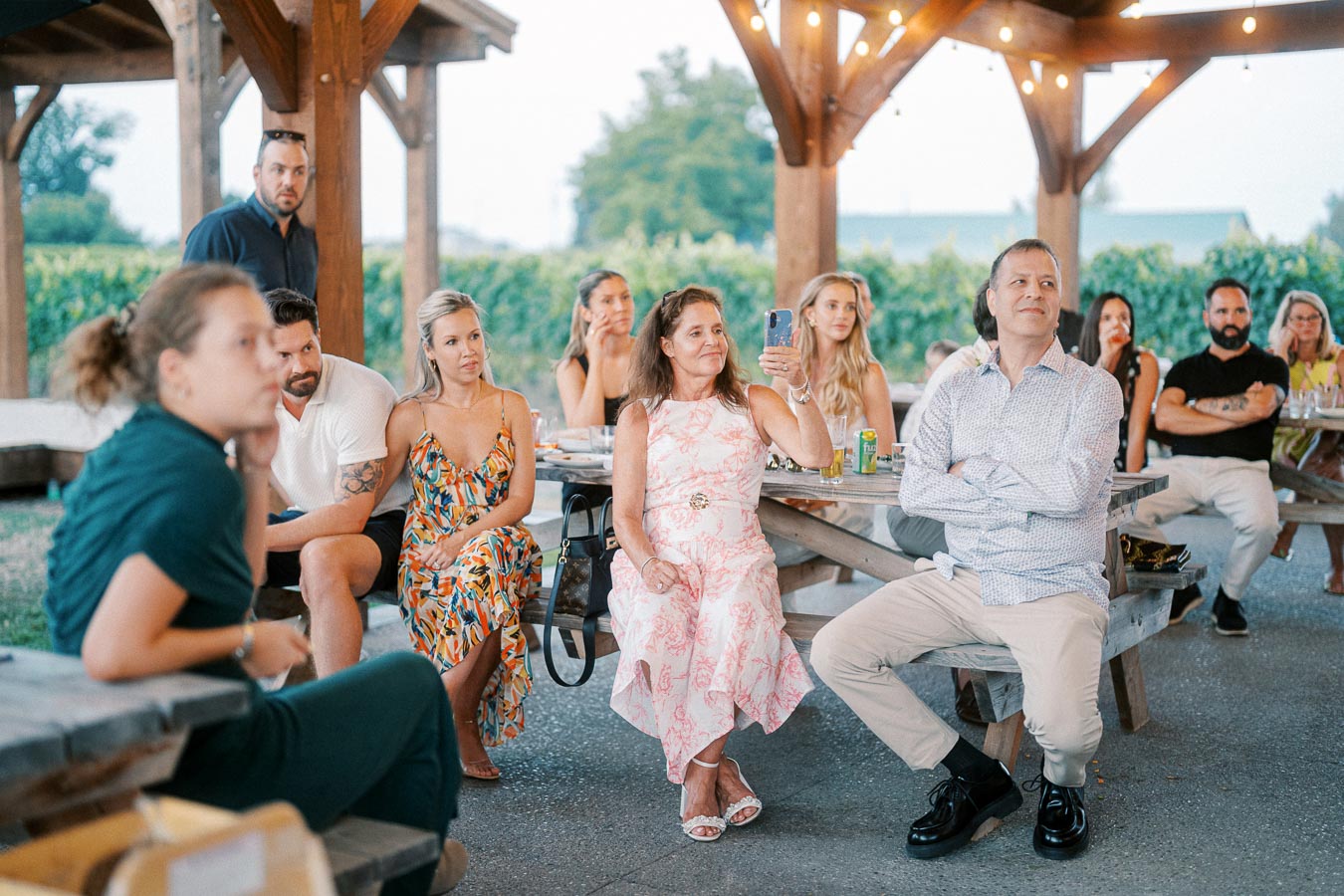 Group of people sitting together under a wooden pavilion at an outdoor event, attentively listening, with festive string lights and green foliage in the background.