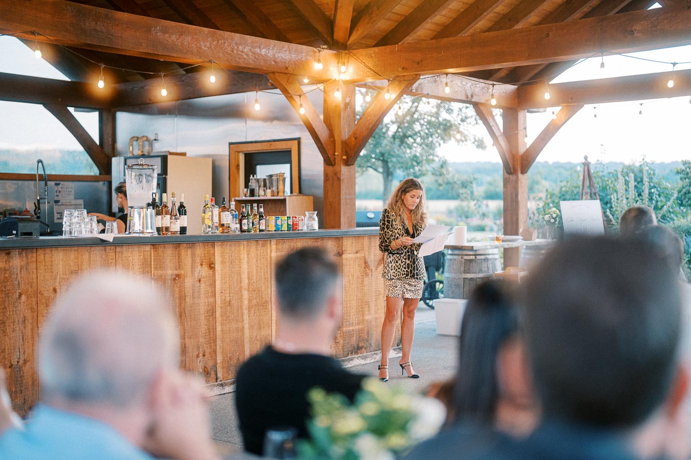 A woman in a leopard print outfit stands at a rustic wooden bar reading from papers during an outdoor event with hanging string lights and a scenic view. Bottles and drinks are displayed on the bar counter, and attendees are seated in the foreground.