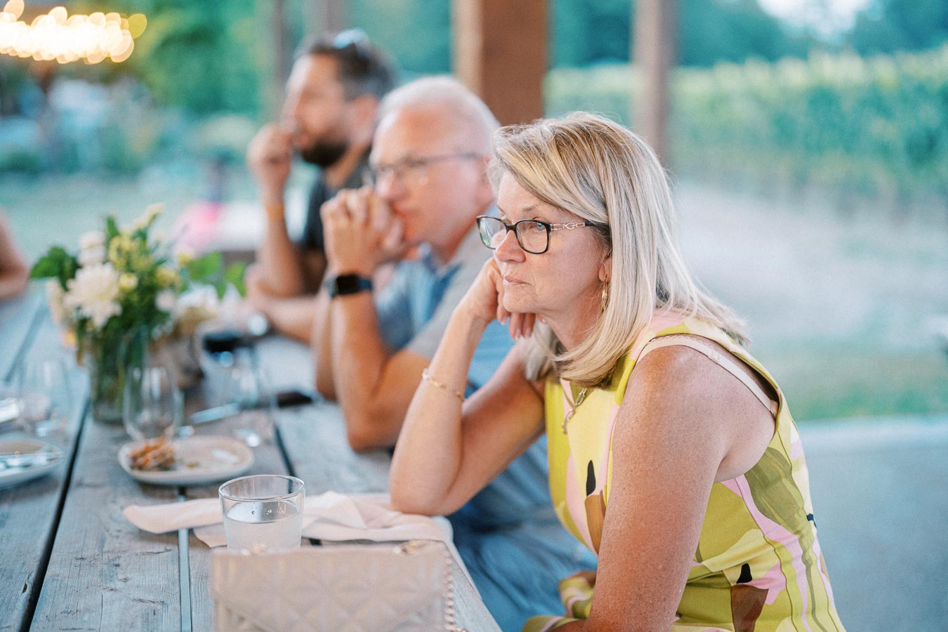 Women and men sitting at an outdoor wooden table during a social gathering, with blurred background and decorative lighting.
