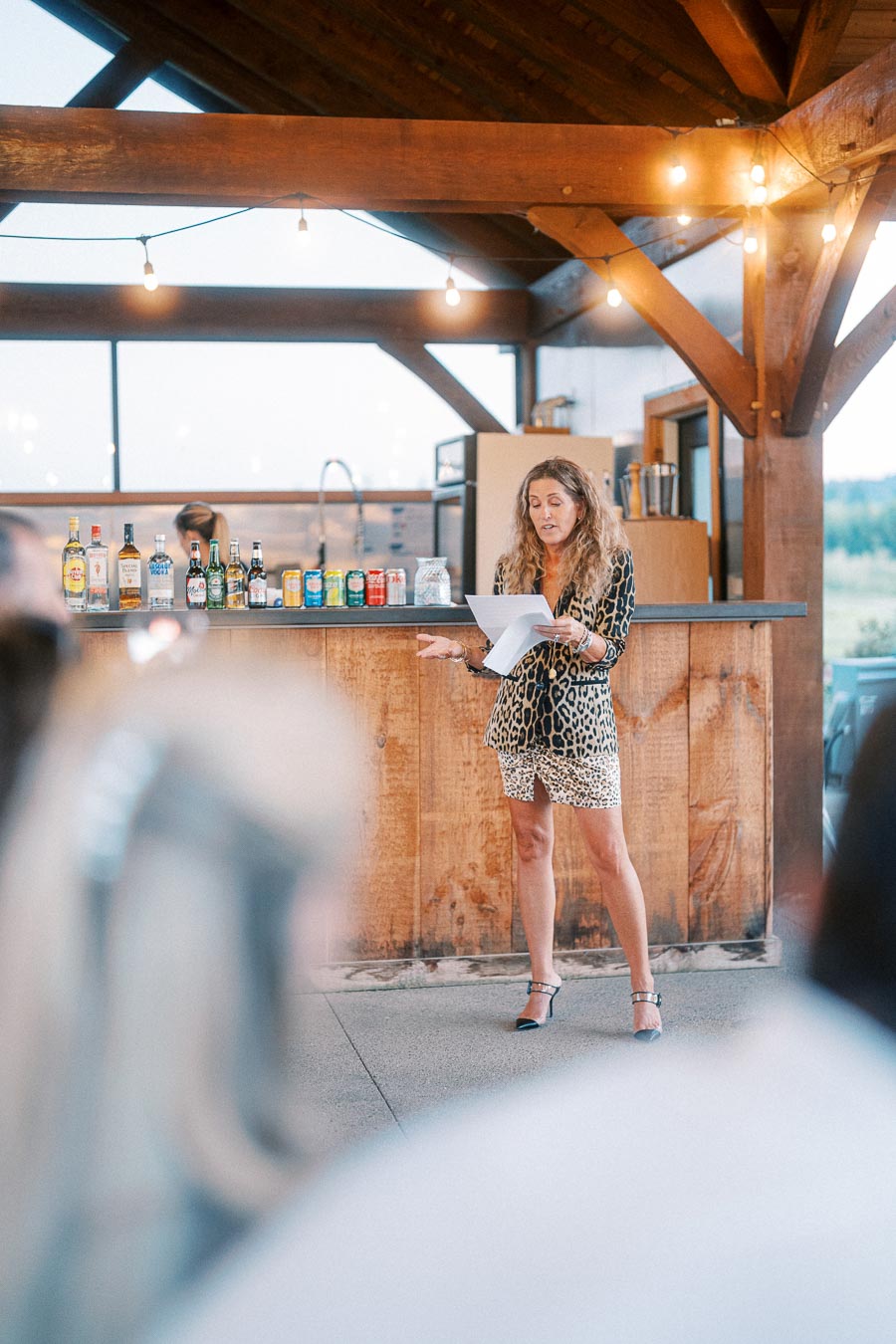 Woman in a leopard-print outfit giving a speech at a wooden outdoor bar area with assorted drinks visible in the background.