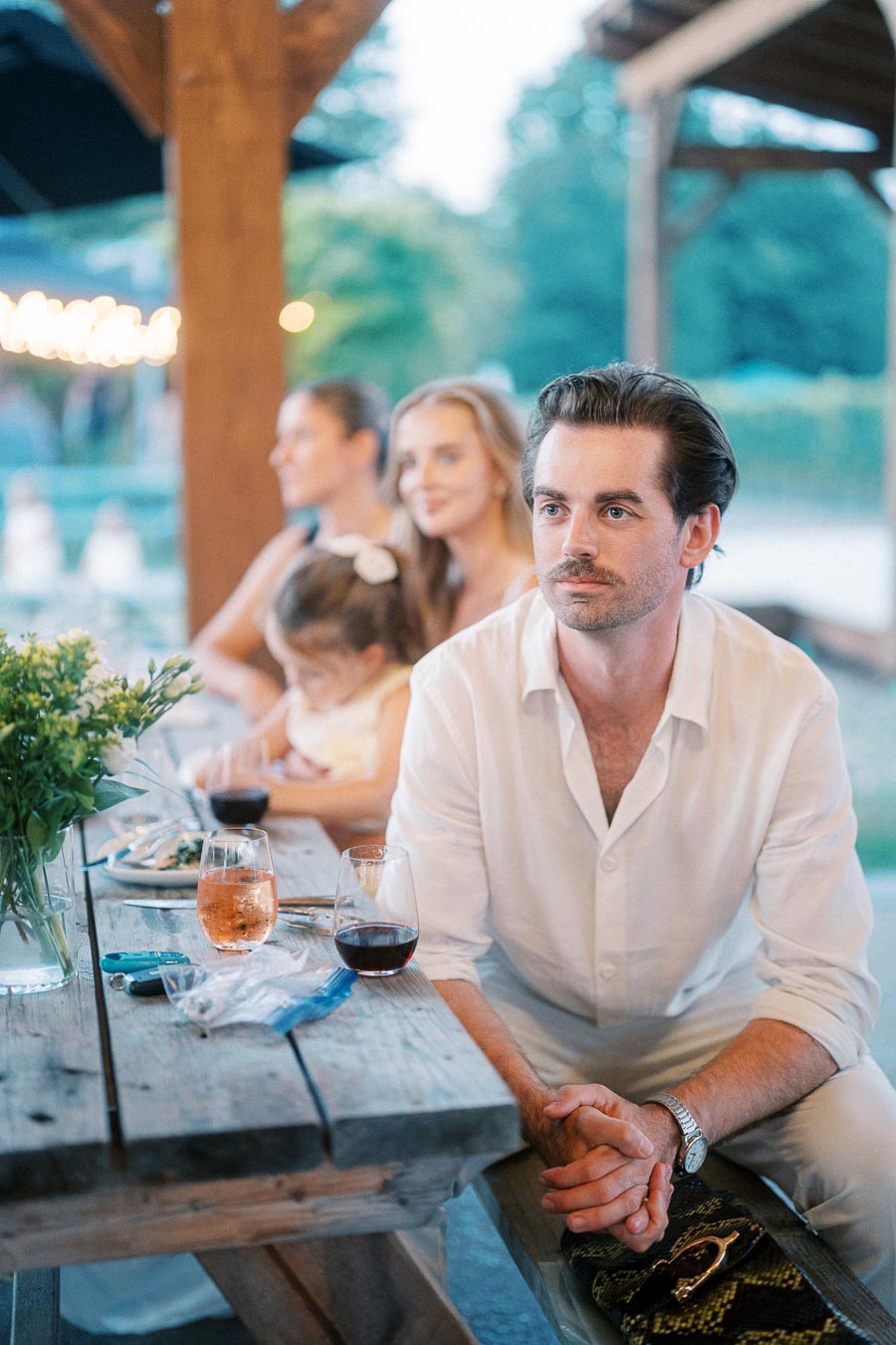 A group of people seated outdoors at a rustic wooden picnic table, enjoying a casual gathering. The focus is on a man in a white shirt looking thoughtfully into the distance, with glasses of wine and a floral arrangement visible on the table, suggesting a warm and social evening setting.