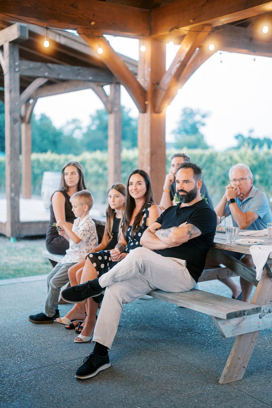 A family sits together at an outdoor picnic table under a wooden pavilion, surrounded by lush greenery, with string lights creating a warm and inviting atmosphere.