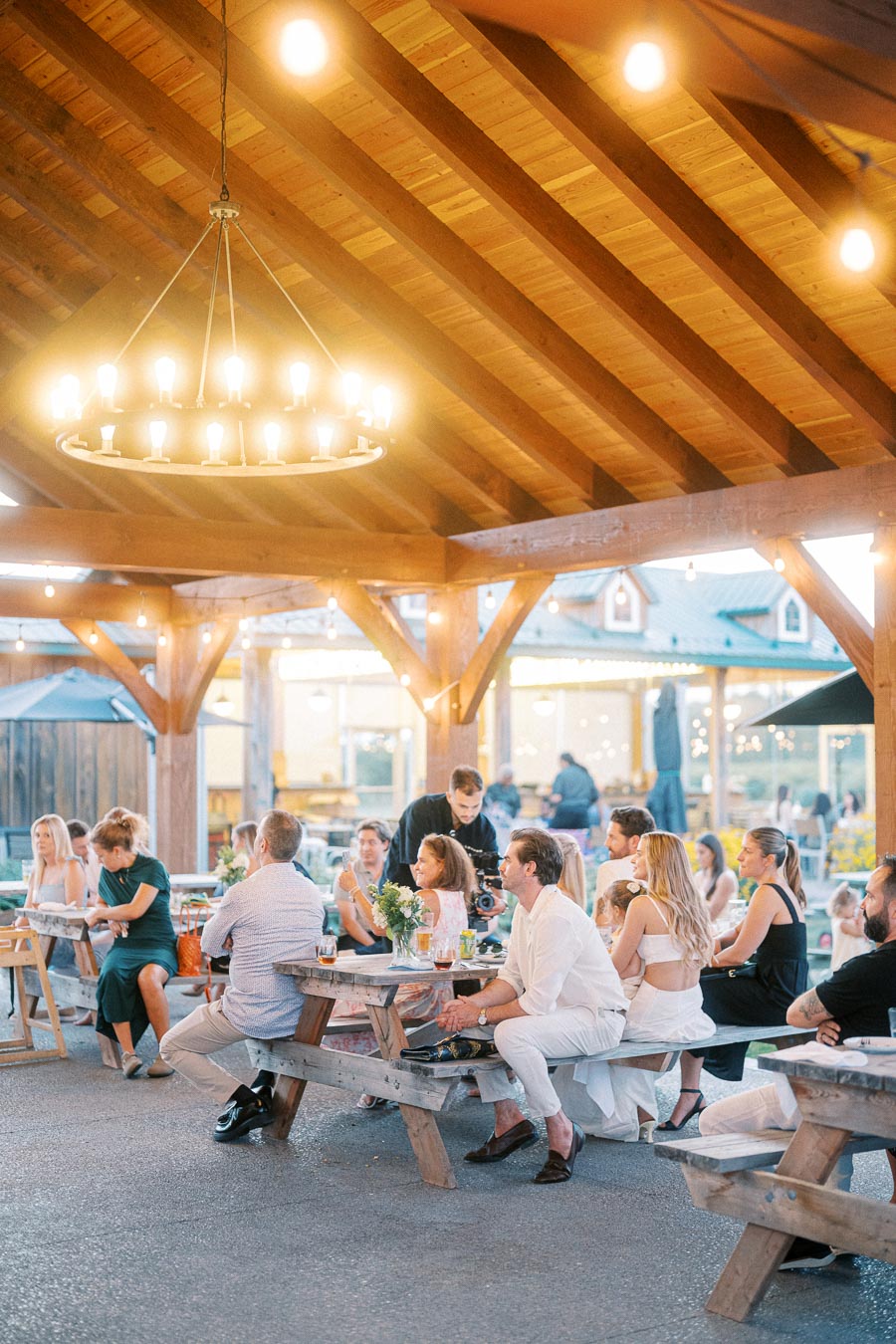 Group of people enjoying an outdoor gathering under a wooden pavilion with warm lighting, seated at picnic tables, surrounded by a relaxed atmosphere.