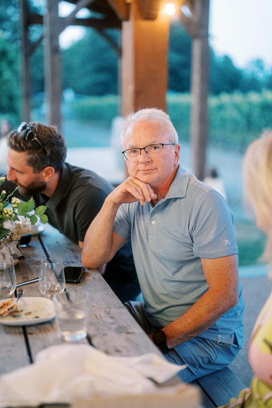 A contemplative man sitting at a rustic outdoor table, wearing glasses and a blue polo shirt, with a blurred background of trees and soft lighting, creating a serene and thoughtful atmosphere at a social gathering.