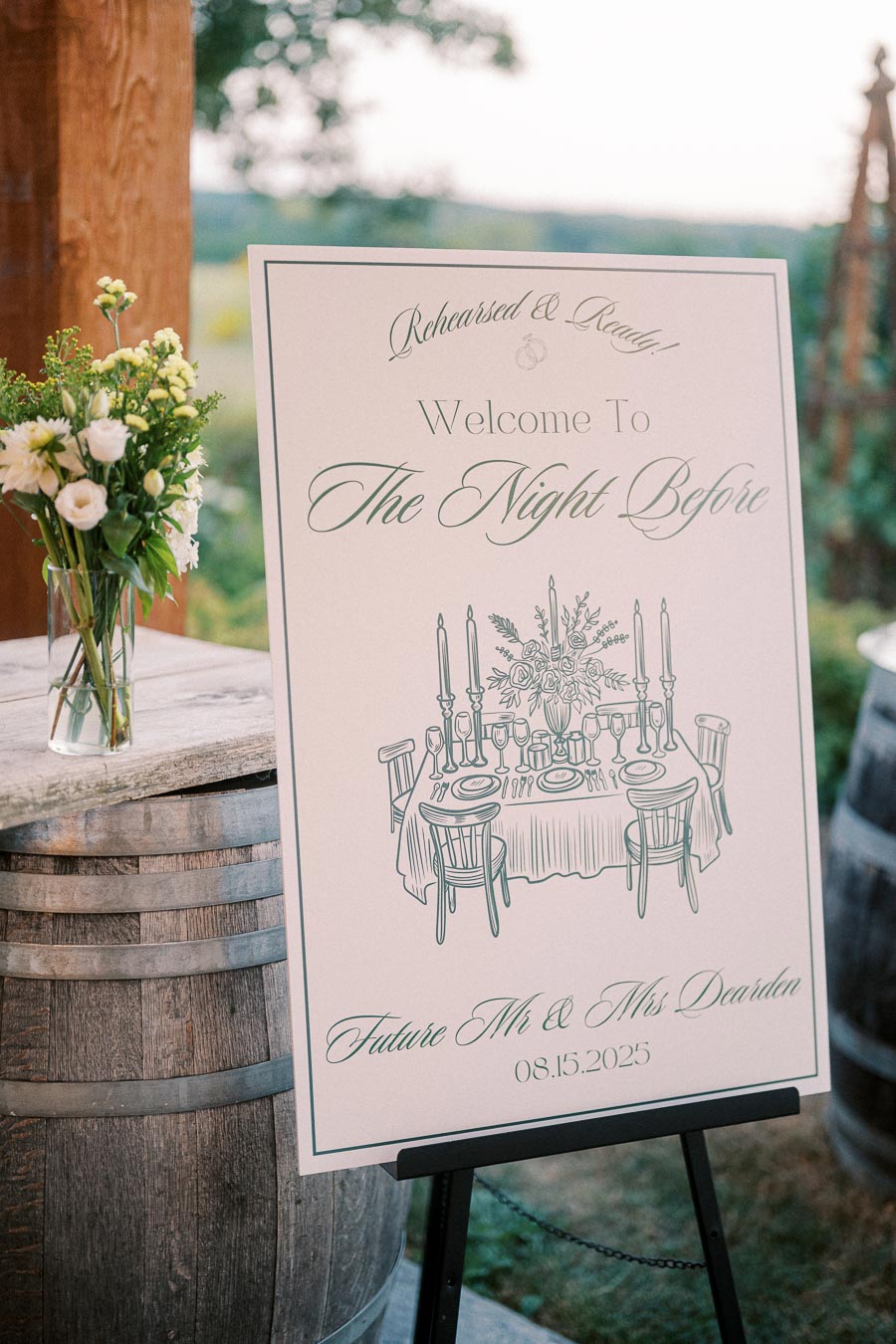 Wedding rehearsal dinner sign with elegant typography and illustration of a table setting, placed outdoors near rustic wooden barrels and a small floral arrangement, welcoming guests to The Night Before event for the future Mr. and Mrs. Dearden, dated 08.15.2025.