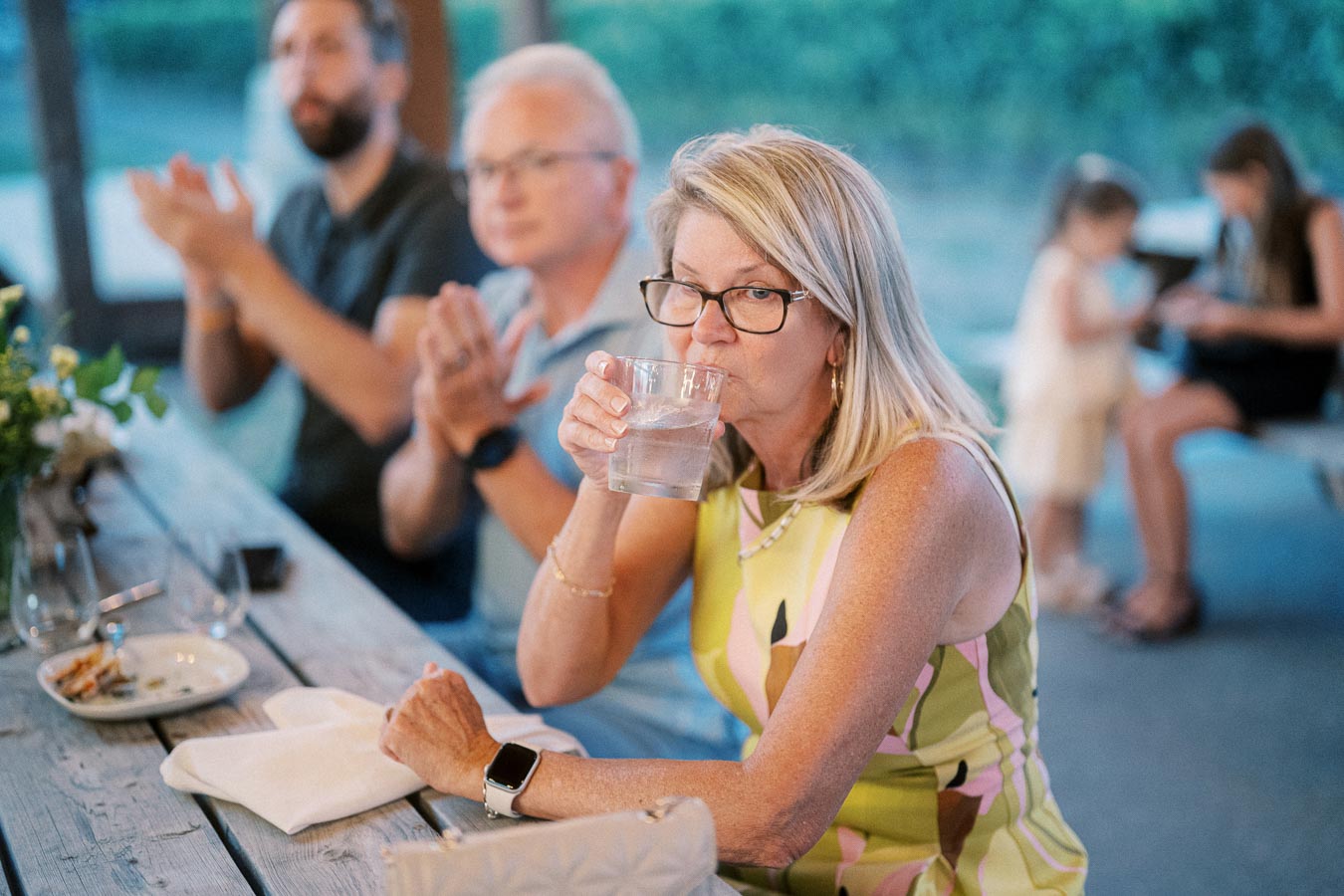 A woman in a colorful dress drinks from a glass at a social gathering, seated at a wooden outdoor table. She is surrounded by blurred attendees clapping, with a casual atmosphere and greenery in the background.