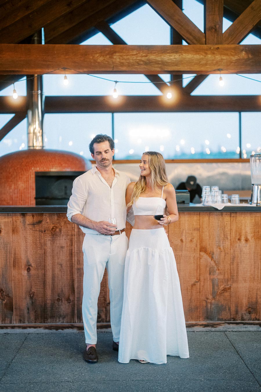 A couple in white outfits standing by a rustic wooden bar indoors, holding glasses of wine, with soft ambient lighting and a brick oven in the background.
