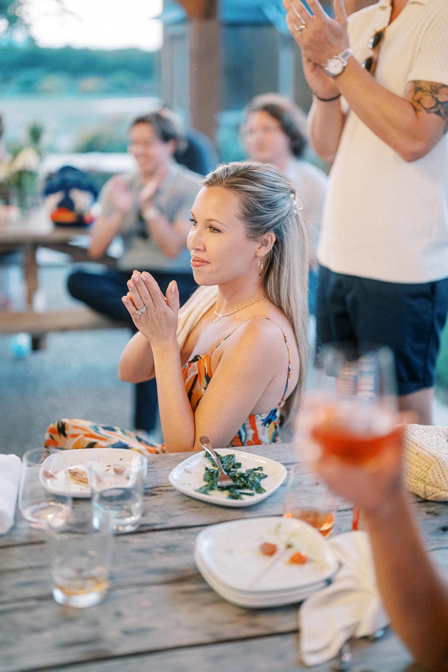 A woman in a colorful dress sits at a wooden table during an outdoor gathering, surrounded by people clapping. Plates with food remnants and drinks are on the table, creating a lively, social atmosphere.