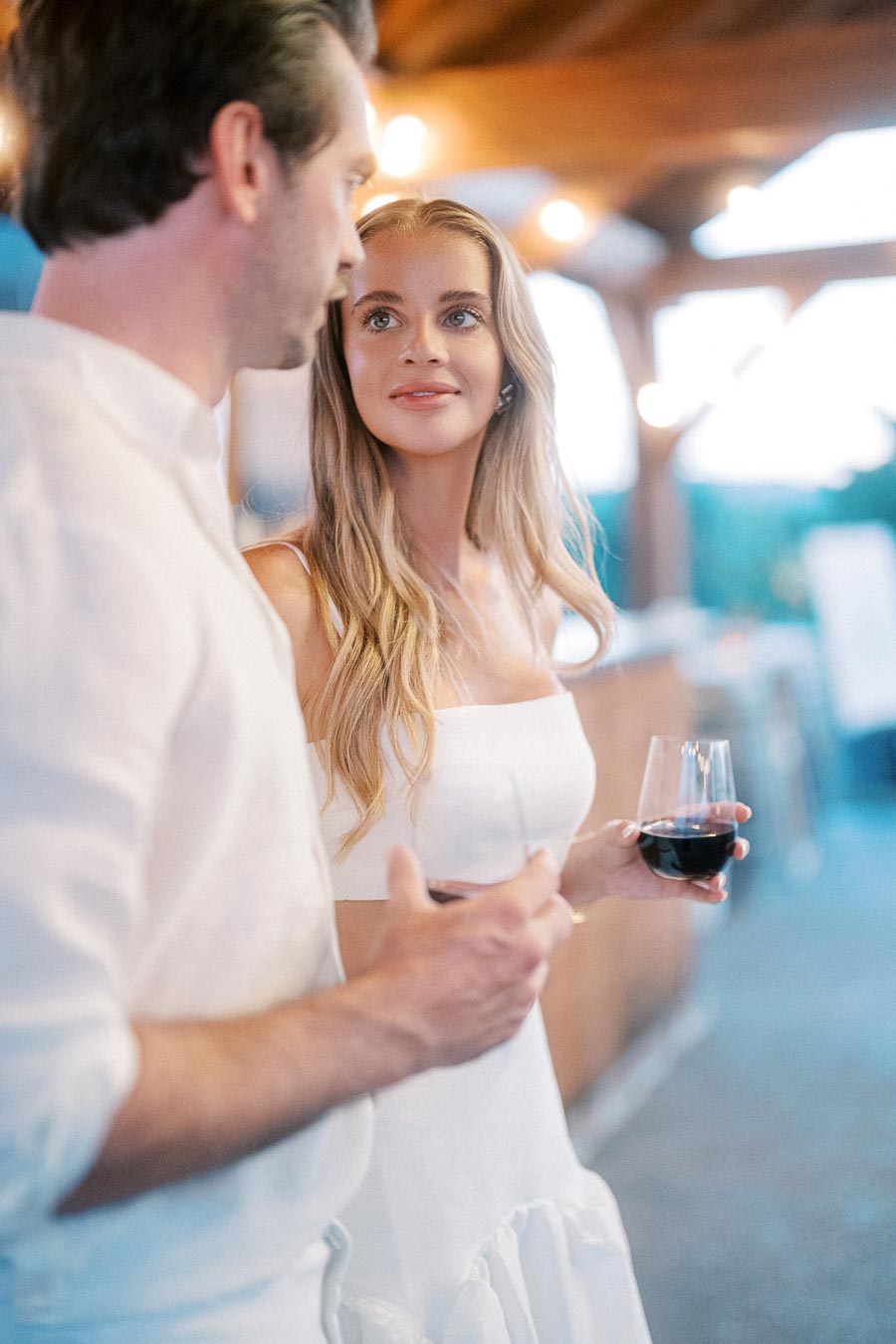 A couple enjoying a romantic evening outdoors, dressed in white attire, holding glasses of red wine under warm ambient lighting.