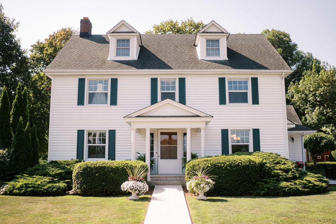 White two-story colonial-style house with green shutters and a manicured lawn, featuring a central front door with steps leading to a path lined with decorative shrubs and flowers.
