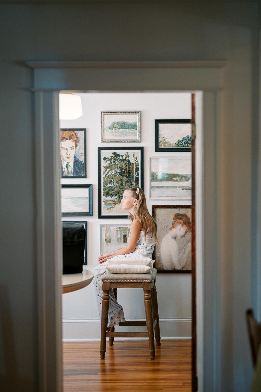 Girl sitting on a stool in an art-filled room, surrounded by framed paintings on the wall, with natural light illuminating the space.