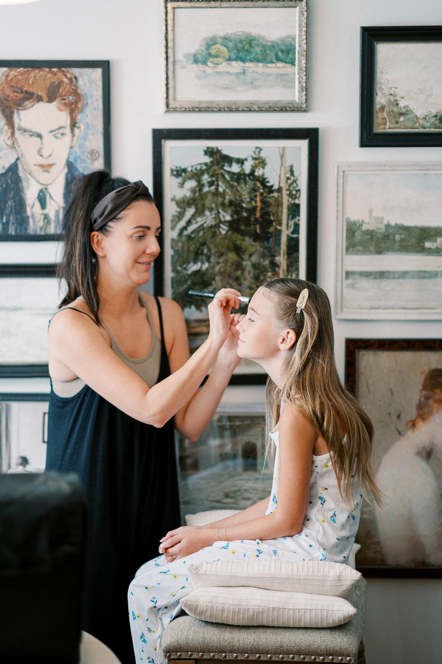 Woman applying makeup to a young girl in an art-filled room.