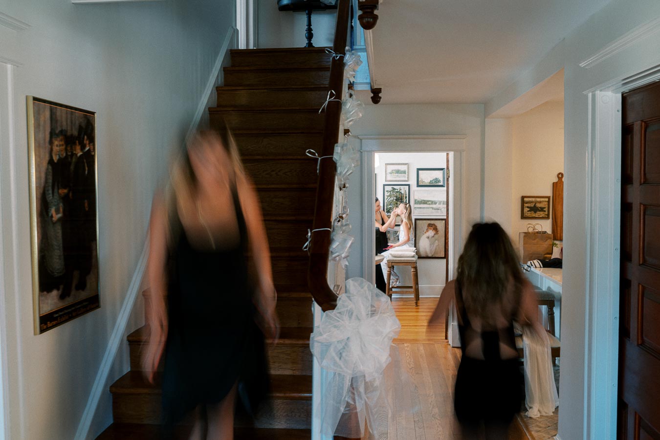 A busy hallway setting with a staircase on the left, decorated with white tulle. Two blurred figures in black dresses move in the foreground. In the background, a person is getting their makeup done by a makeup artist, surrounded by framed artwork in a well-lit room.