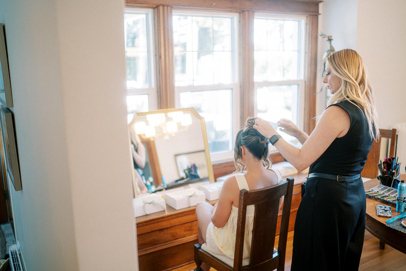 A woman styling another woman's hair in a sunlit room with a large window and a wooden dressing table.