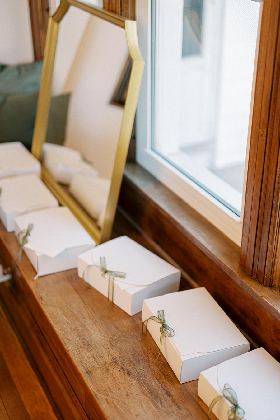 White gift boxes tied with green ribbons lined up on a wooden ledge next to a window and mirror.