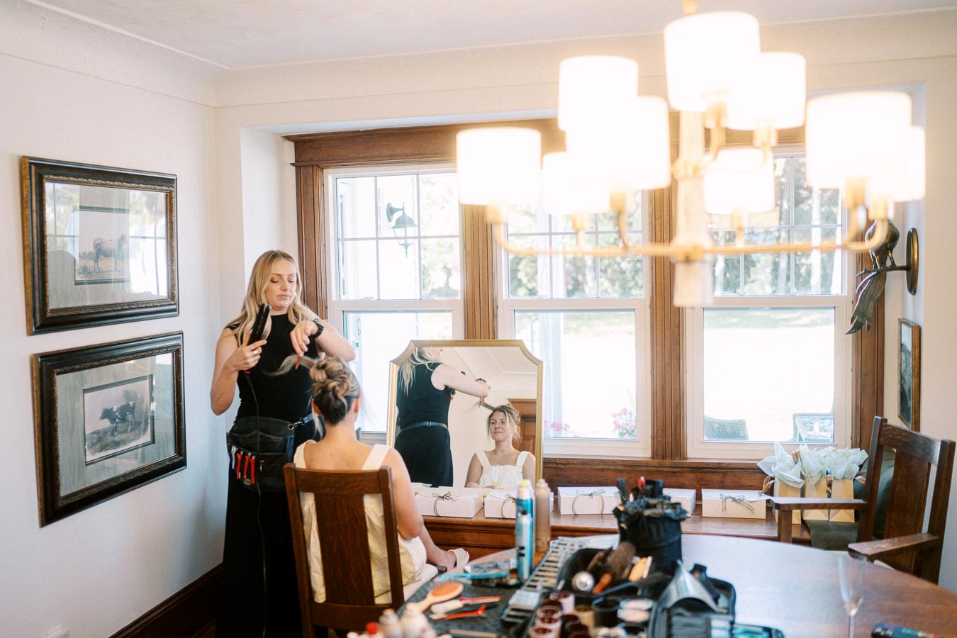 A hairstylist works on an elegant bridal updo for a young woman seated in front of a large mirror within a warmly lit room, with hairstyling tools and products arranged on the table.