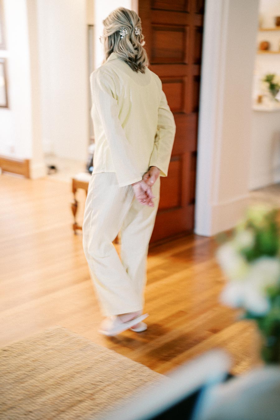 Elderly woman in light-colored pajamas with hair clips walking indoors on polished wooden floor.