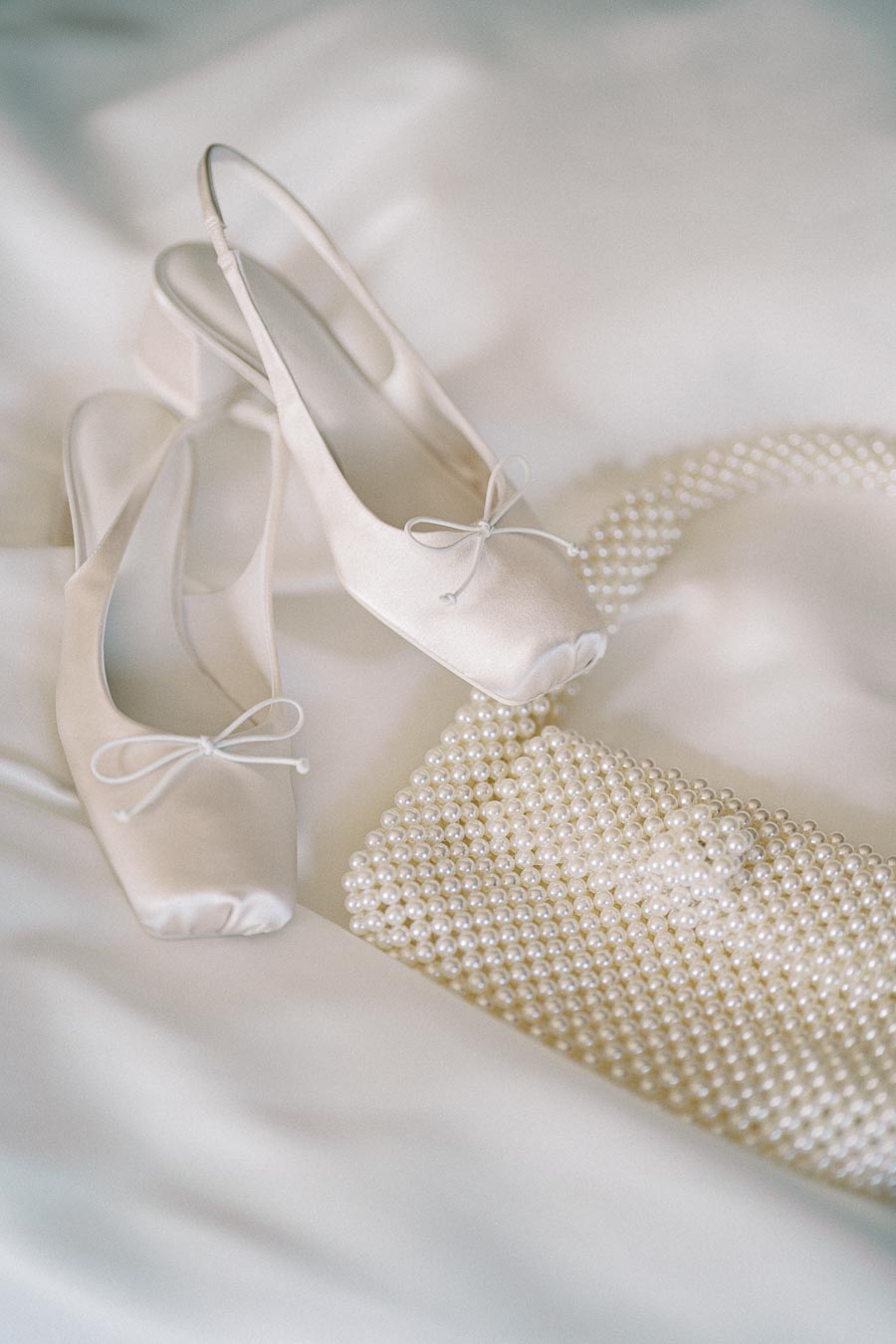 Elegant cream satin bridal shoes with bows paired with a pearl-embellished clutch on a soft, white fabric background. Ideal wedding accessory ensemble.